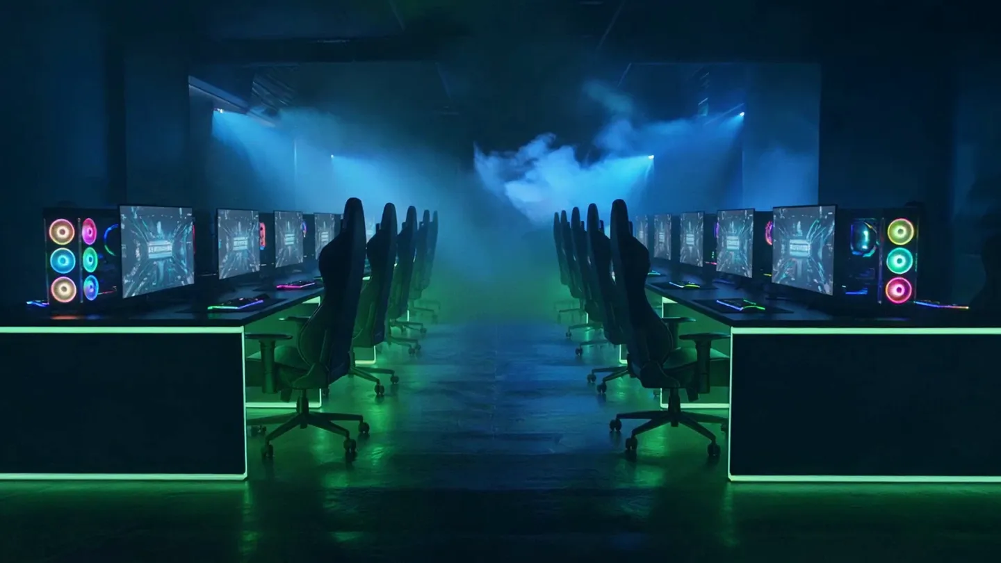 Tamsi žaidimų salė su kompiuterių eilėmis ir žalia šviesa ant grindų