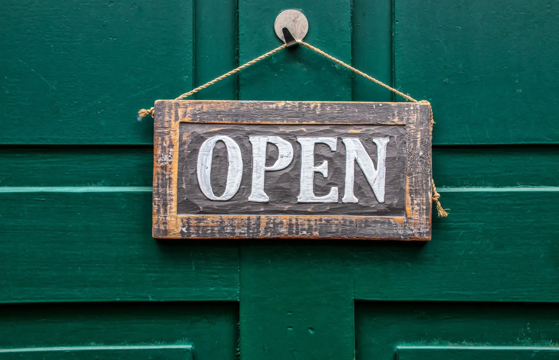 Medinė lentelė su užrašu „Open“, pakabinta ant žalių durų